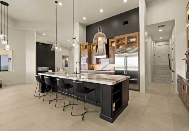 A spacious kitchen featuring a large dark island with a marble countertop, black bar stools, and pendant lighting. The background includes a stainless steel refrigerator, range hood, and wooden cabinets, with a hallway leading to upper levels visible in the distance.